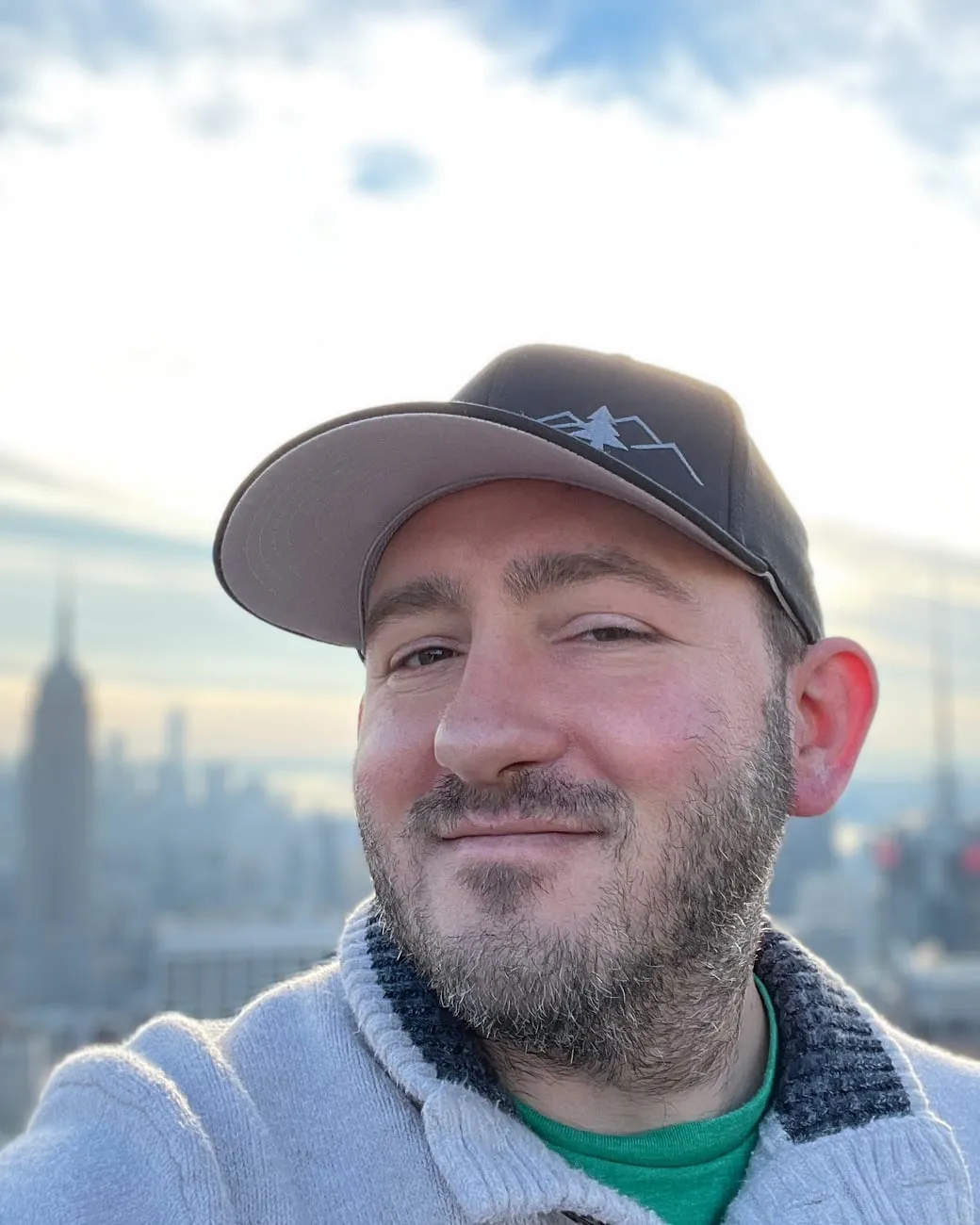 Joe Henzi in a grey cap and sweater, smiling, with the Manhattan skyline and the Empire State Building soft behind him