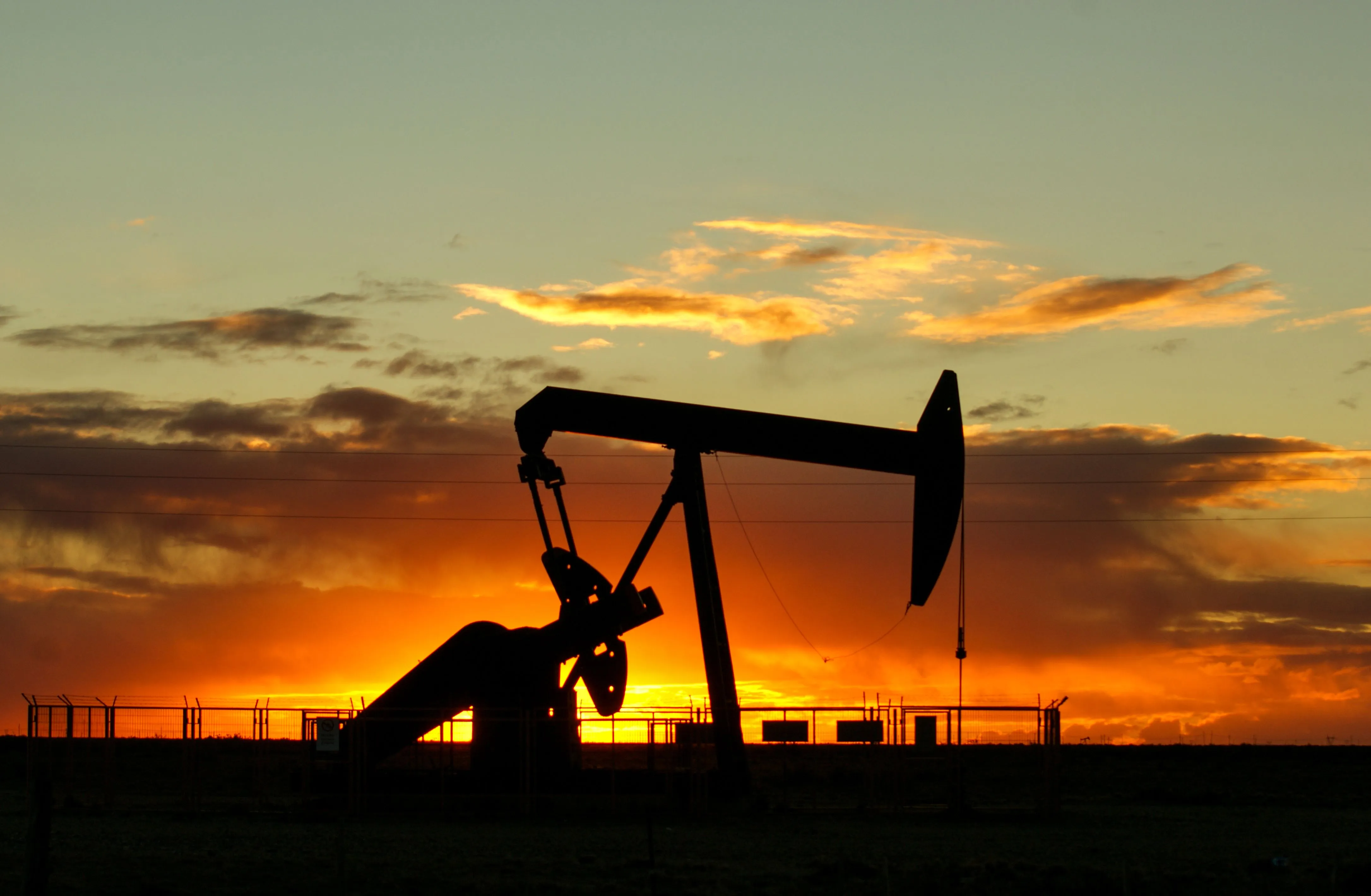 An oil pump jack in silhouette against an orange sunset, its horsehead tipped down toward the well. A chain-link fence runs behind it across flat, dark ground, power lines cross the sky, and streaks of cloud glow gold above.